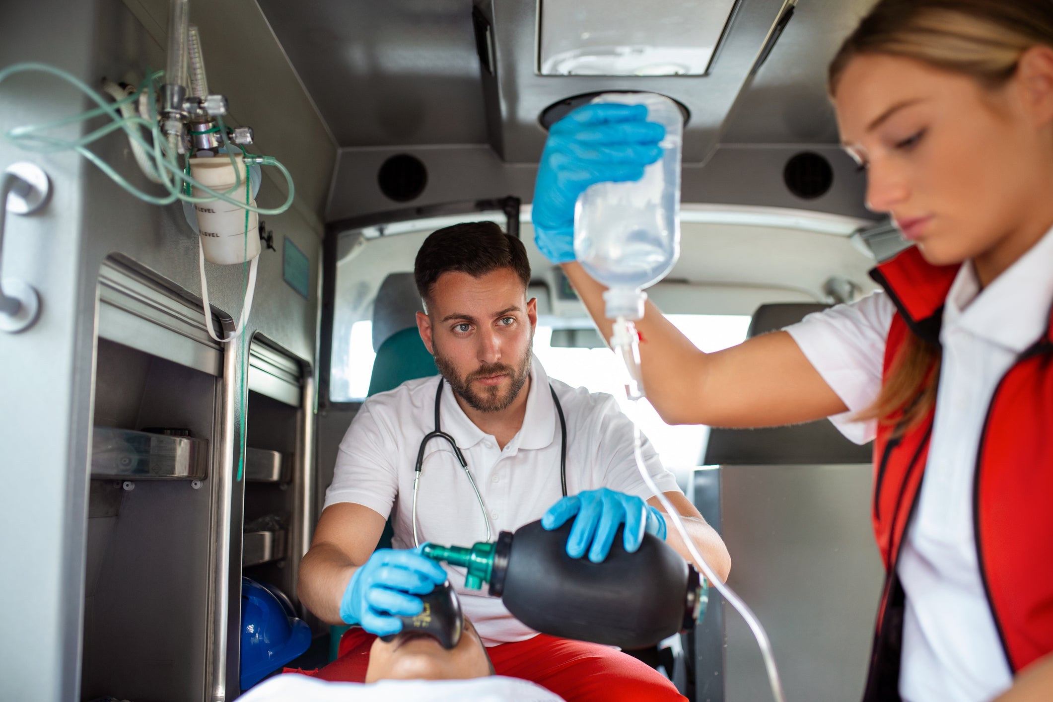 Two emergency responders inside an ambulance preparing medical equipment, with one holding a bag and the other using a manual pump. The scene is set in a real environment with medical supplies visible.