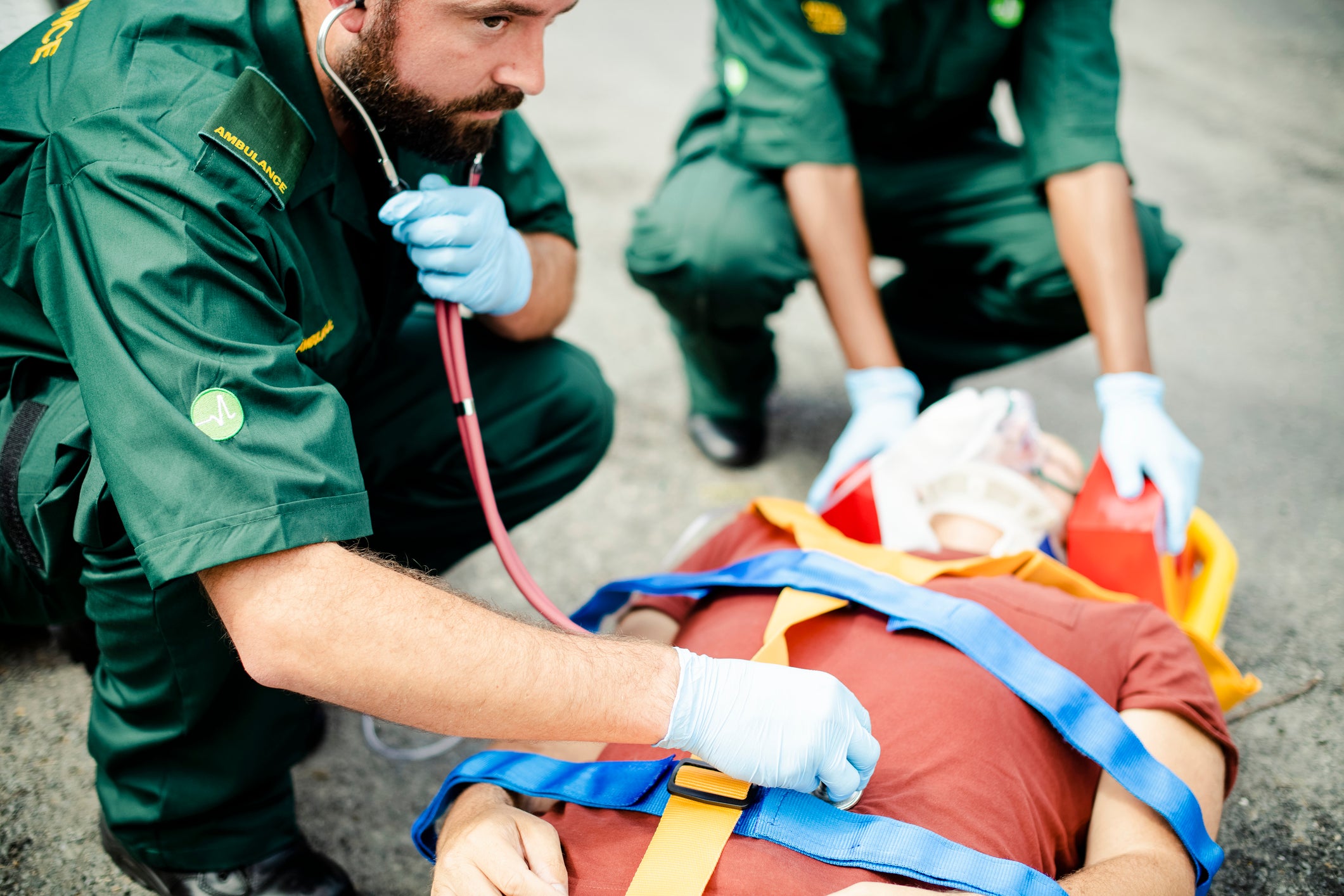 Emergency responders in green uniforms attending to a person on a stretcher, with one responder using a stethoscope and another applying an oxygen mask in an outdoor setting.