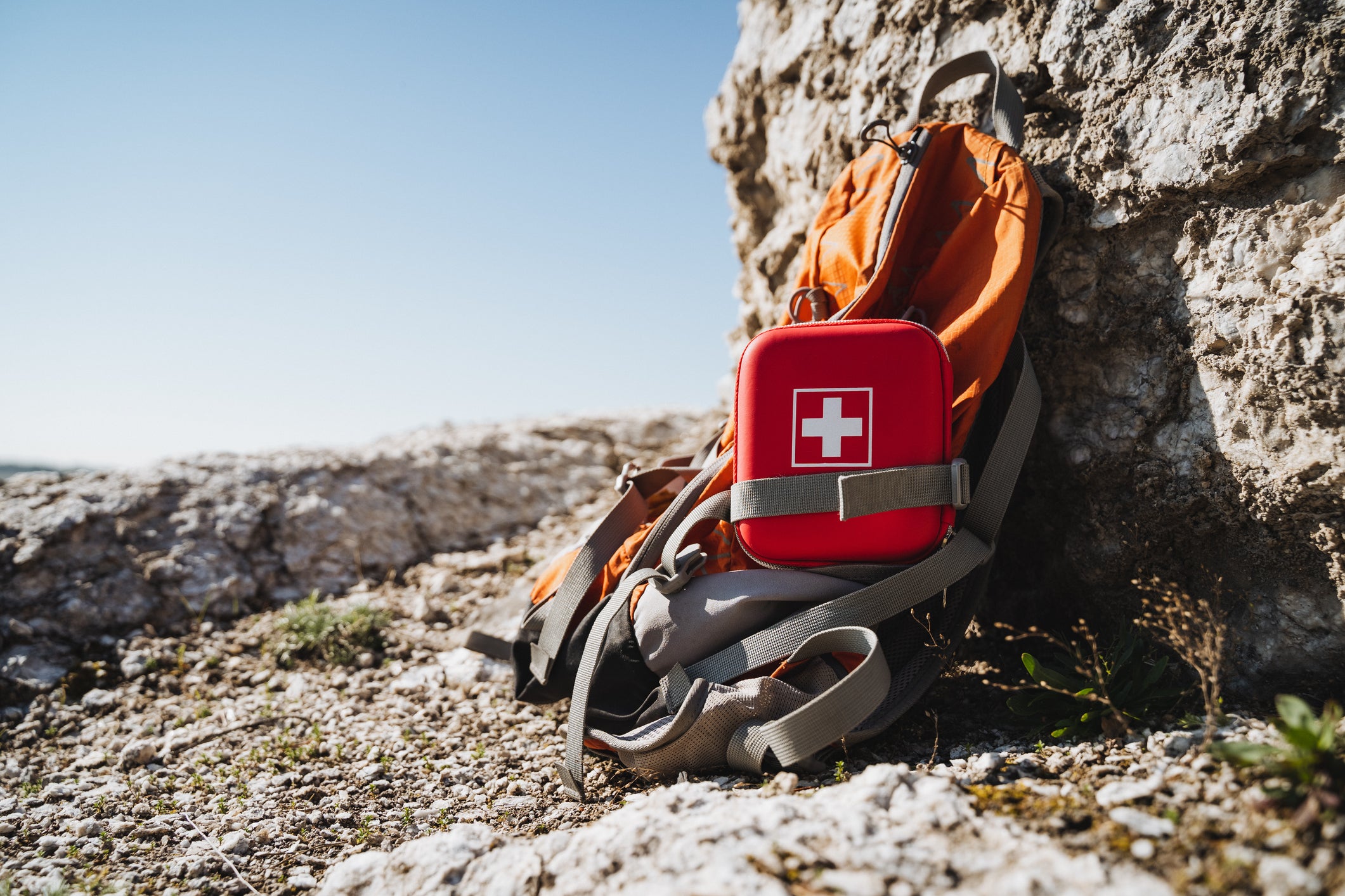 An orange hiking backpack and a red first aid kit with a white cross are placed on rocky ground against a large stone wall under a clear blue sky.