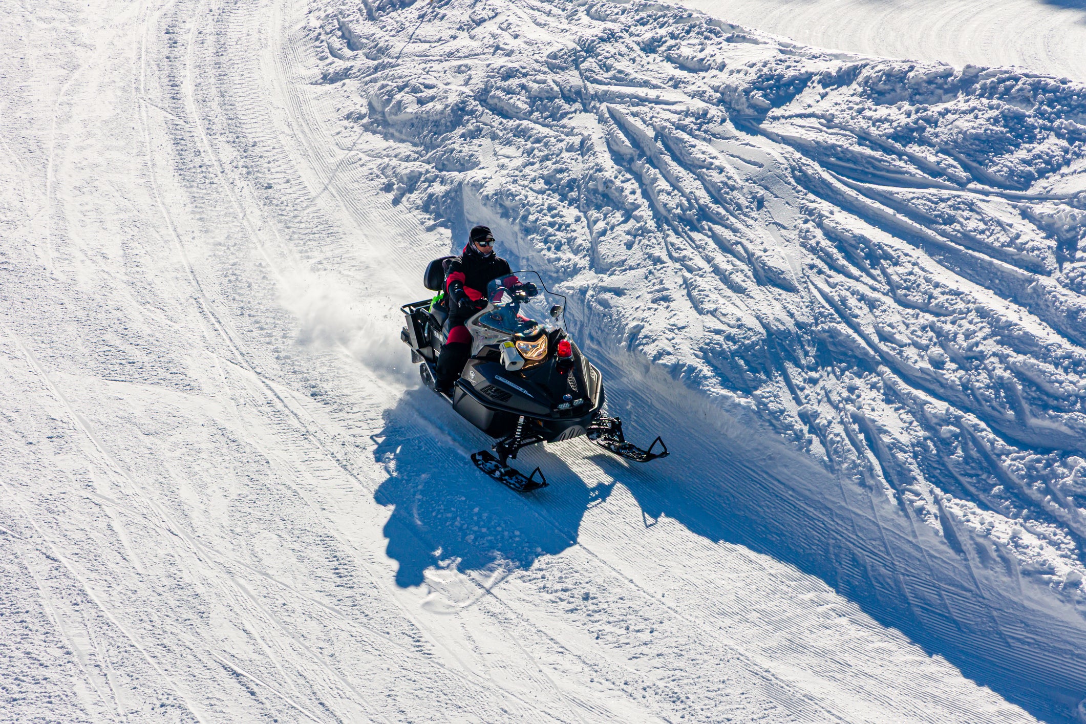 A snowmobile with two riders moves through deep snow tracks on a snowy mountain slope, creating a spray of powder and casting strong shadows.