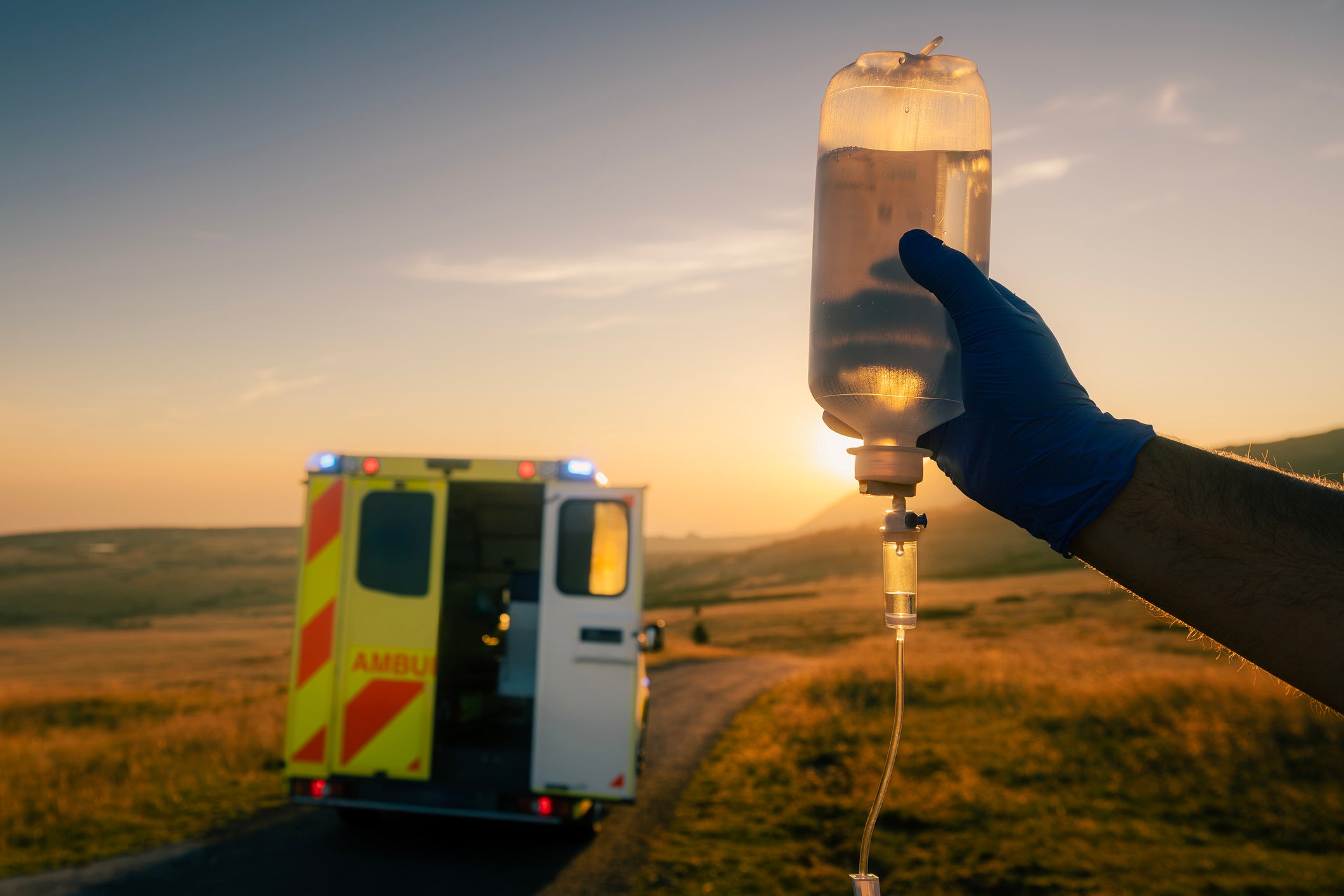 A gloved hand holds an IV bag in the foreground at sunset, with an ambulance parked on a road in the blurred background.