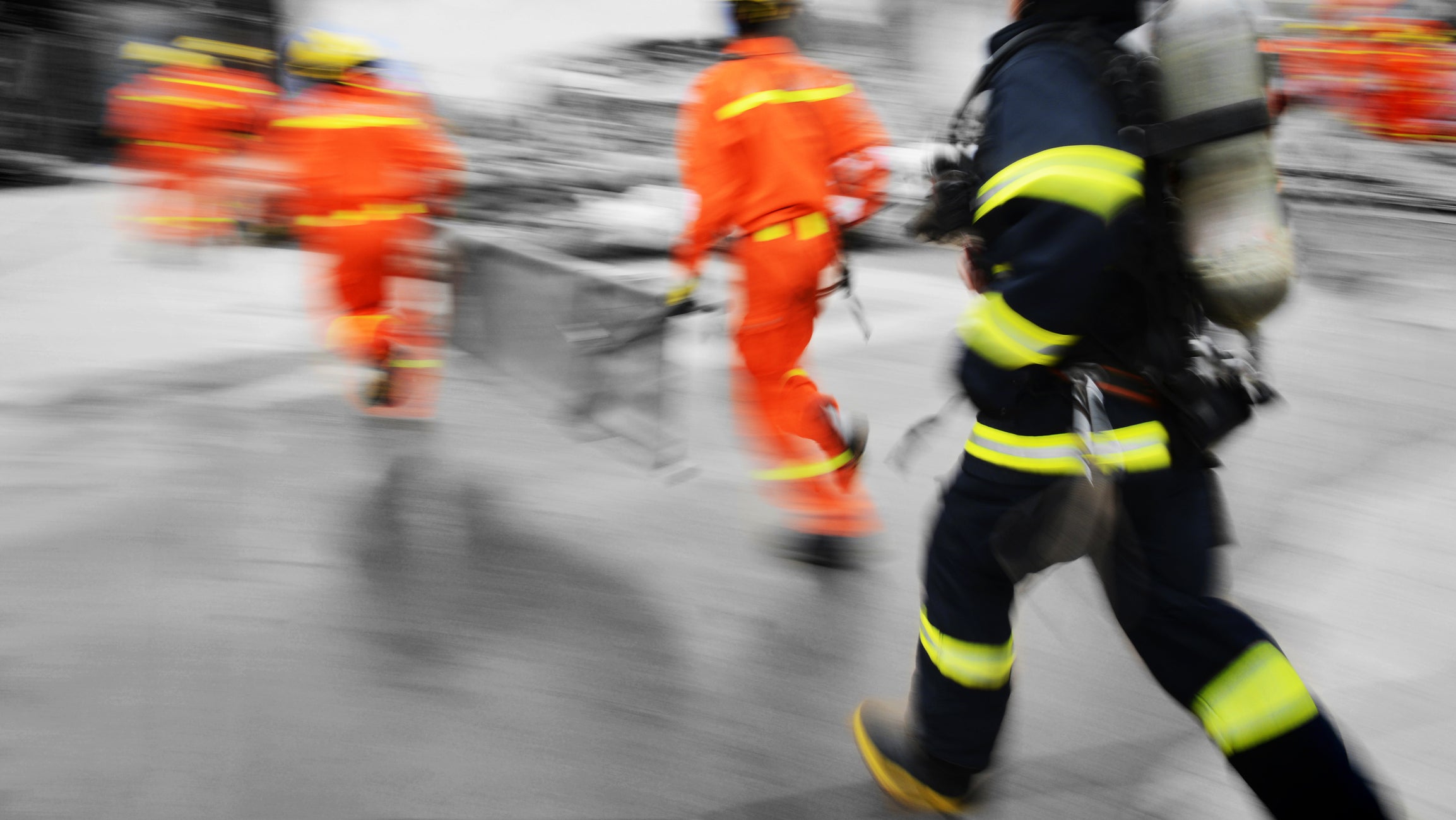A dynamic, motion-blurred action shot of firefighters in action, featuring a firefighter in the foreground wearing a dark navy uniform with bright yellow reflective stripes and carrying an oxygen tank, alongside other crew members in orange turnout gear in the background.